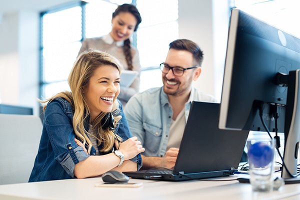 Three people smile; two are sitting at a desk in the foreground in front of two computers, and one is in the background holding paperwork.