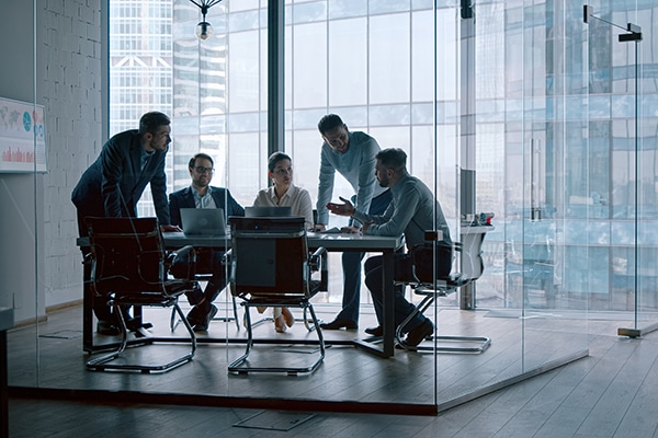 people sitting and standing around a table in an office, holding a meeting